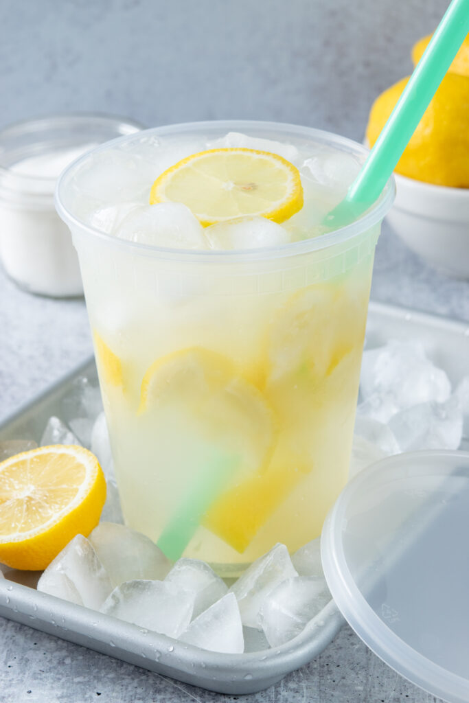 A refreshing state fair lemonade drink with a green straw in it, on a small tray that's filled with iced cubes and half a lemon. Next to the drink, which is in a 32oz deli container, is a lid, a small bowl of sugar and a bowl with lemons in it.