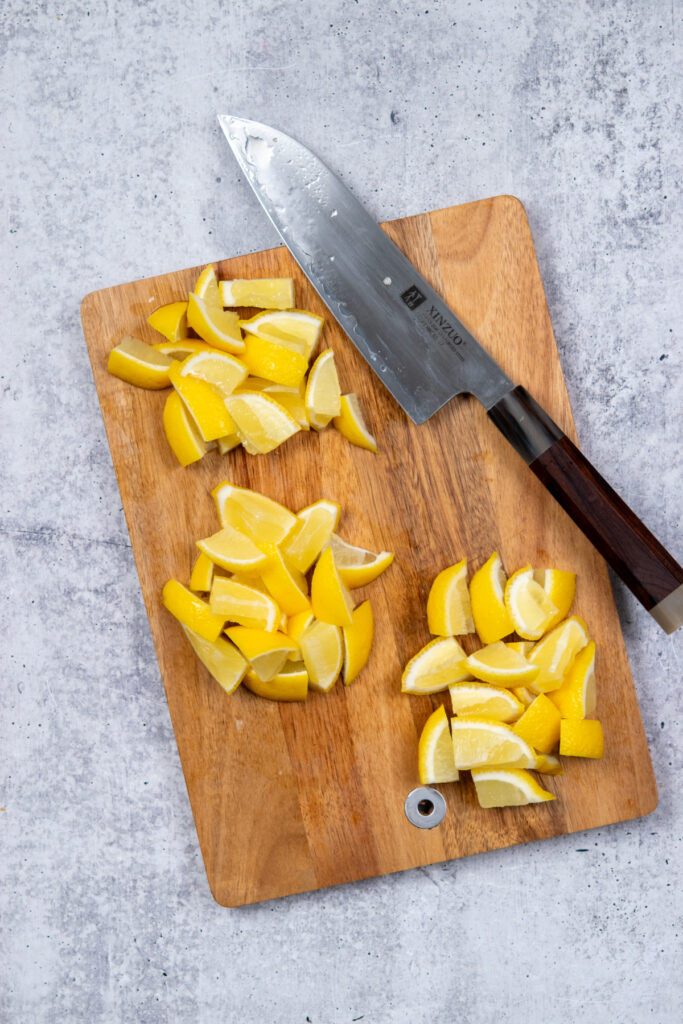 An overhead photo showing how to cut three whole lemons into wedges and chunks for smashing into homemade state fair lemonade. The piles of lemons are on a cutting board next to a knife.