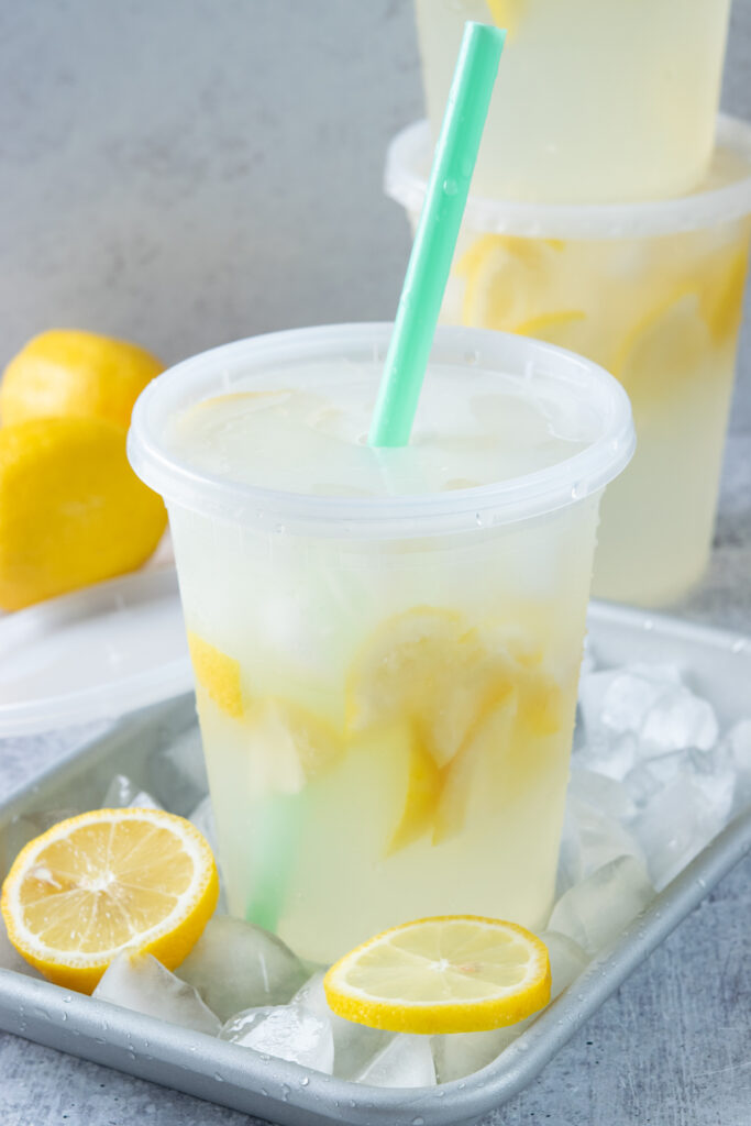 A homemade state fair lemonade made in a 32 ounce deli container with lid that has a hole punched in it for  a straw, just like how it's served on food trucks. The lemonade is on a tray covered in ice and there are lemons and two more lemonades in the background. The drinks look icy and refreshing.