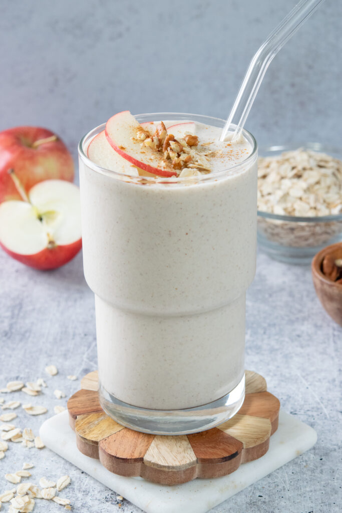 An apple cinnamon smoothie with thins slices of gala apple and cinnamon on top. In the background is an apple, half an apple and a small bowl of oats.