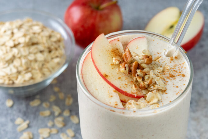 An apple smoothie surrounded by a bowl of oats and red apples. There are thin slices of red apple, cinnamon, oats and chopped pecans on top of the breakfast smoothie.