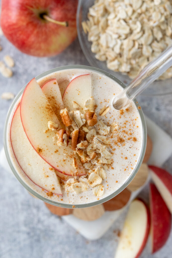 An overhead, close-up view of an apple cinnamon smoothie. There's a glass straw in the drink and the to is garnished with slices of gala apple, chopped pecans, oats and cinnamon. It looks thick and creamy.