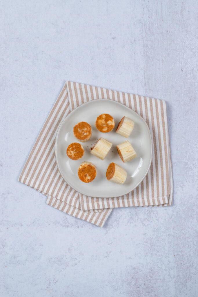 A banana cut into one-inch chunks in a single layer on a tray to be frozen for a smoothie. Each piece of banana is dipped in cinnamon so it's ready to add to the freezer pack, along with the other dry ingredients: chia seeds and powdered peanut butter.