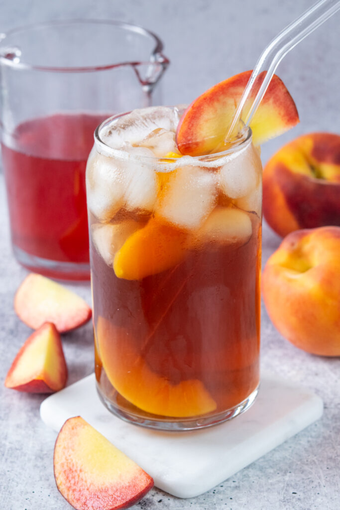 Glass of peach iced tea garnished with a peach wedge and peaches inside, with a bottle of homemade peach syrup and a fresh peaches in the background.
