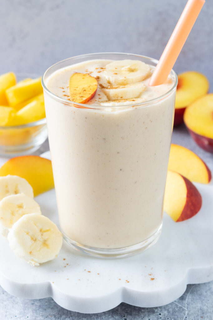 The creamiest peach and banana smoothie in a cup that has an orange straw in it. The smoothie is surrounded by banana and peach slices.
