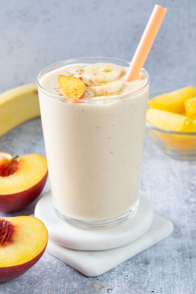 A peach and banana smoothie in a tall glass. The smoothie is topped with three slices of banana, a slice of peach, and cinnamon sprinkled on top. There's a banana and bowl of peaches in the background.