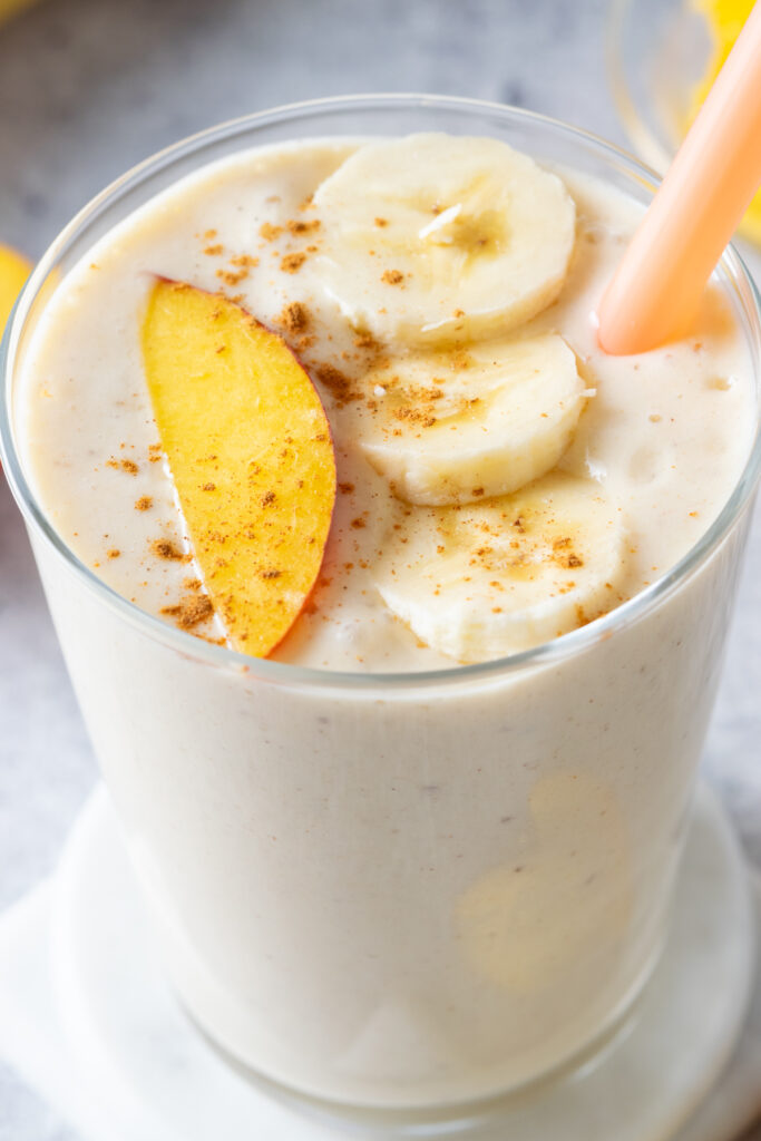 A top-down photo of a creamy peach and banana smoothie. On top of the smoothie is a fresh peach slice and three slices of banana. There is cinnamon sprinkled on top.