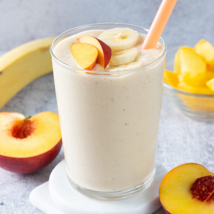 A blended peach banana smoothie in a glass with an orange straw. The smoothie is topped with banana slices and a peach slice. In the background is a full banana and a peach cut in half.