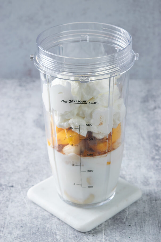 Greek yogurt added to a blender cup on top of cinnamon, fresh peaches, frozen banana and milk.