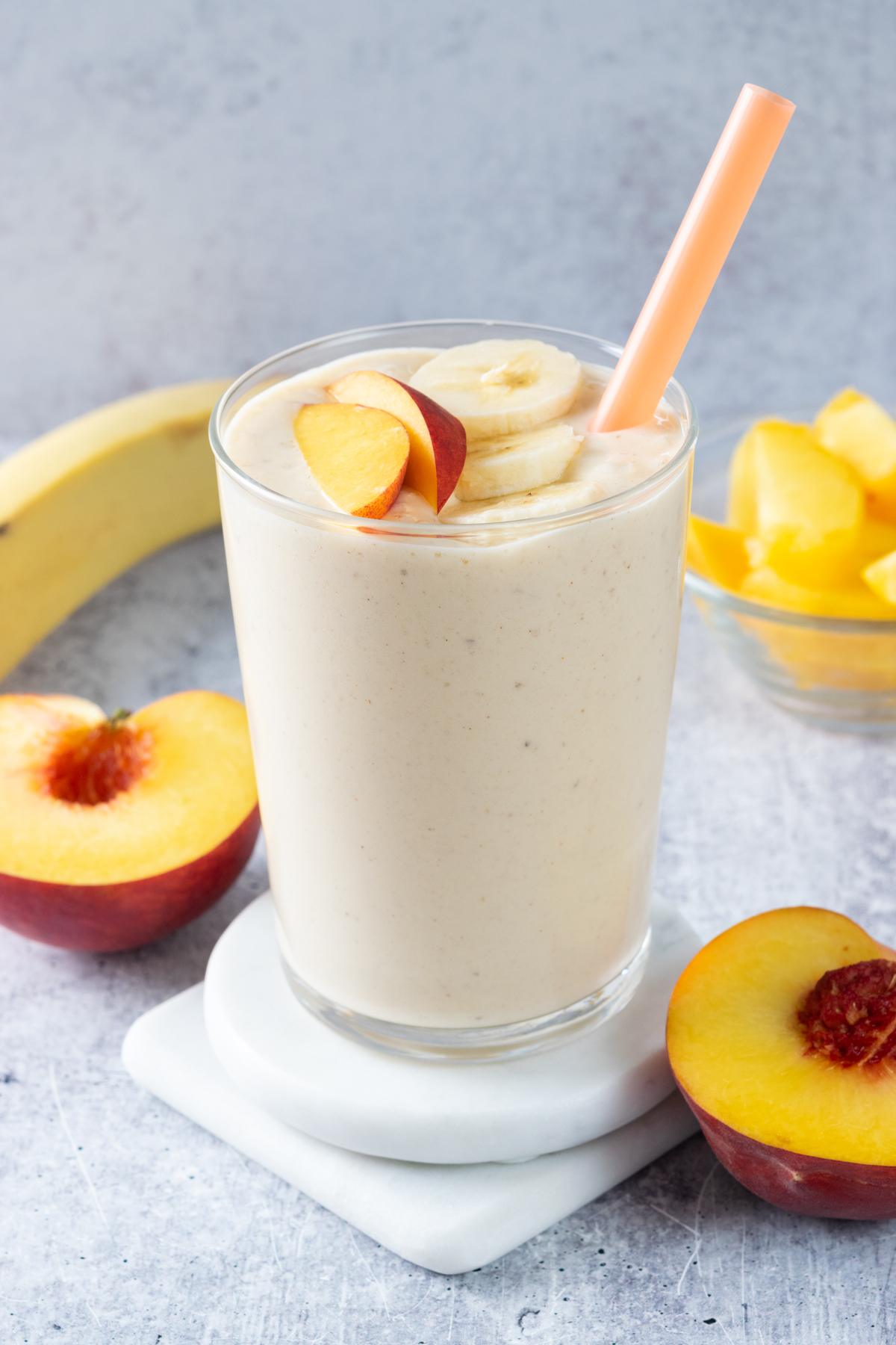 A blended peach banana smoothie in a glass with an orange straw. The smoothie is topped with banana slices and a peach slice. In the background is a full banana and a peach cut in half.