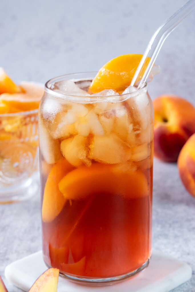Homemade peach iced tea in a tall glass filled with ice. There are peach wedges in the drink and garnished on the rim. The iced tea looks icy, peachy and refreshing.