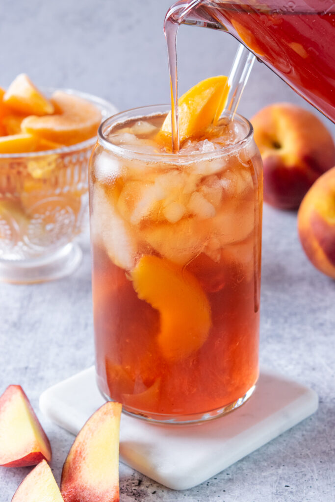 Pouring homemade peach syrup into a glass of iced tea. There are whole peaches and a bowl of sliced peaches in the background.