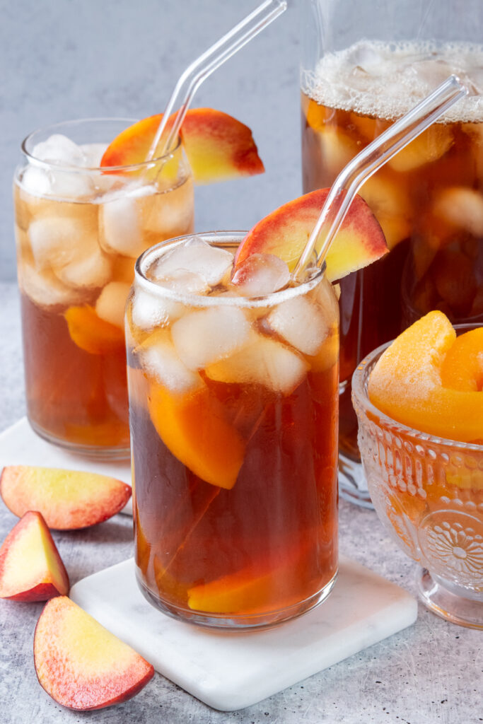 Two glasses of peach iced tea filled with ice and peaches, next to a pitcher of homemade peach iced tea.