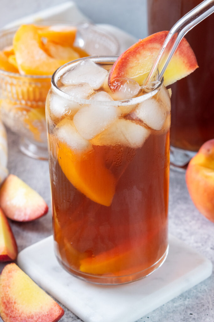 Peach iced tea in a glass filled with ice, and pieces of fresh peach. In the background is a bowl filled with frozen peach wedges for adding to the tea, and a pitcher of peach iced tea.