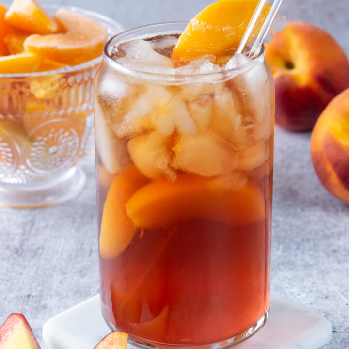 Refreshing glass of homemade peach iced tea. The tea has peaches in it and is garnished with a peach wedge on the rim of the glass. In the background is a bowl of frozen peaches and two whole peaches.