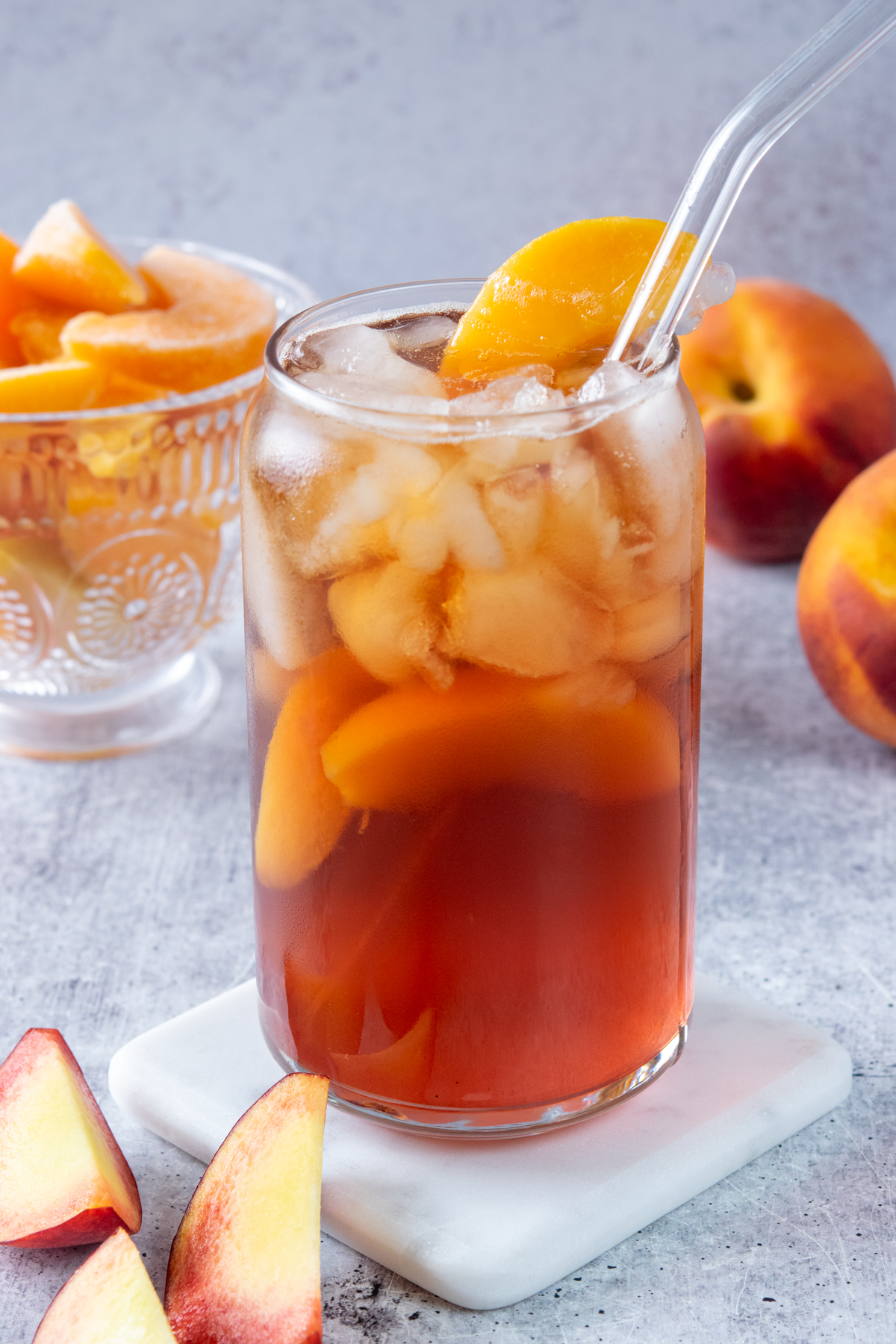 Refreshing glass of homemade peach iced tea. The tea has peaches in it and is garnished with a peach wedge on the rim of the glass. In the background is a bowl of frozen peaches and two whole peaches.