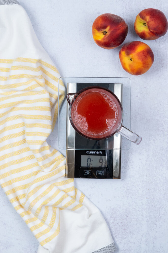 Overhead view of peach juice being weighed on a kitchen scale, with three peaches nearby.