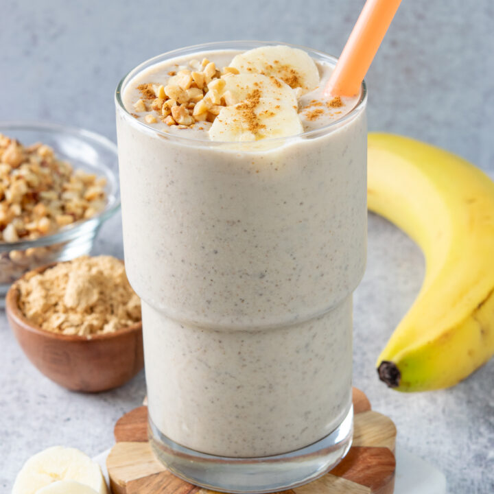 A homemade peanut butter smoothie in a tall glass with an orange straw in it. The thick and creamy smoothie is topped with some crushed peanuts, banana slices and cinnamon. There is a small bowl of powdered peanut butter and a banana in the background.