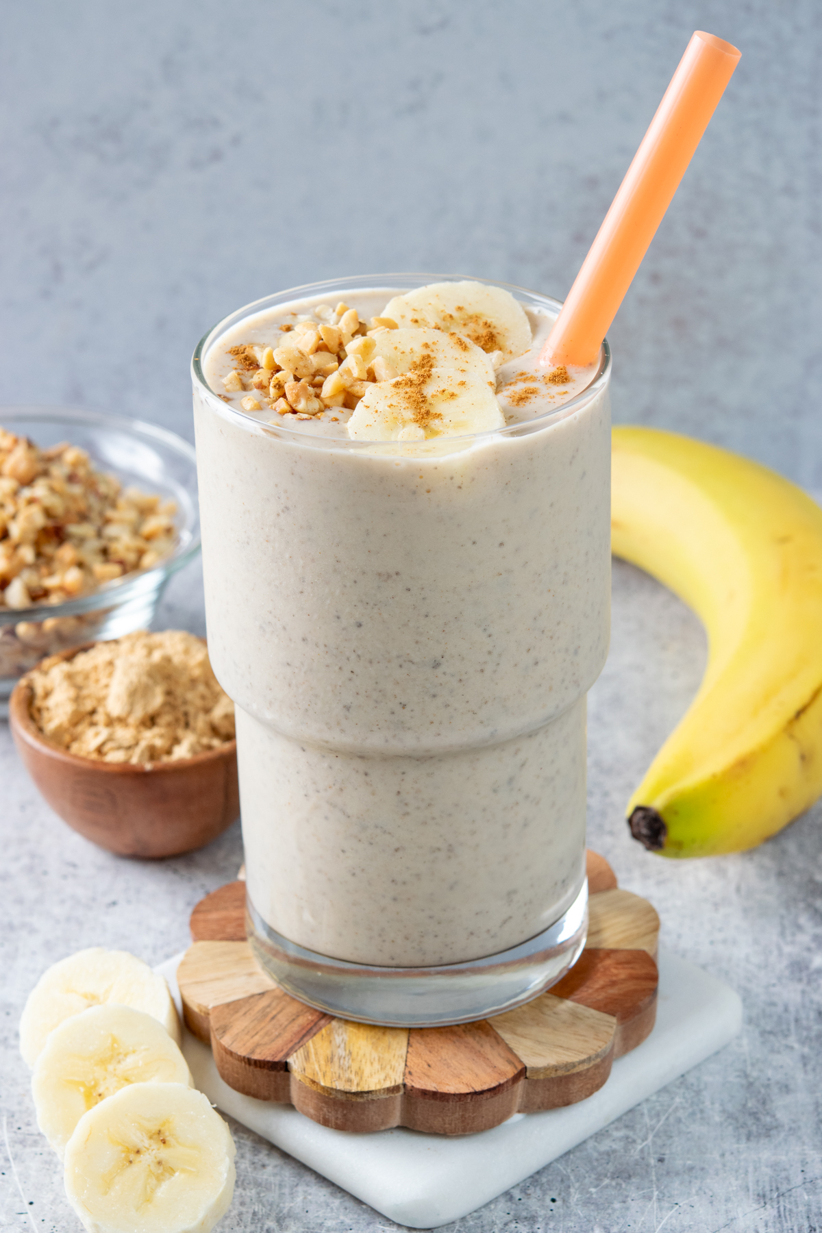 A homemade peanut butter smoothie in a tall glass with an orange straw in it. The thick and creamy smoothie is topped with some crushed peanuts, banana slices and cinnamon. There is a small bowl of powdered peanut butter and a banana in the background.