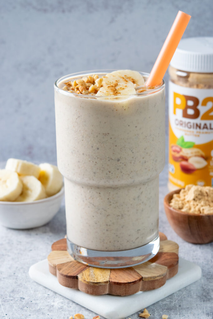 A thick peanut butter smoothie that's filled with protein and bananas. The smoothie is in a tall glass and is topped with peanuts, banana slices and cinnamon. In the background is a jar of powdered peanut butter powder and a small bowl of sliced bananas.