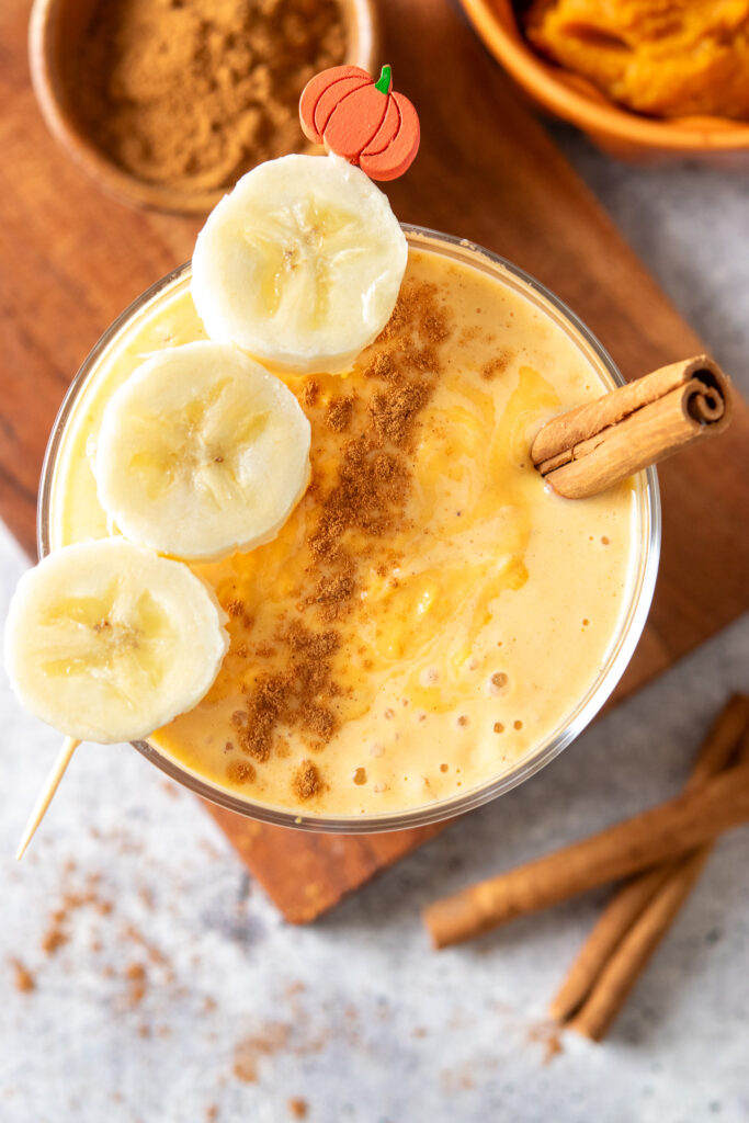The top of a pumpkin smoothie showing that it's thick and creamy and orange-colored. The smoothie has cinnamon sprinkled on top and a cinnamon stick in it. Across the top of the glass is a garnish with 3 slices of banana on a toothpick that has a pumpkin on one end.