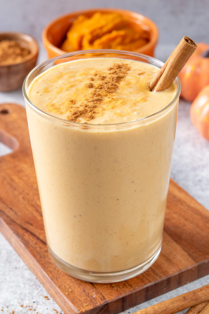 A super creamy, orange-colored pumpkin smoothie in a glass. The smoothie has spice sprinkled on top and a cinnamon stick in it. Next to the smoothie are some mini pumpkins, pumpkin puree in an orange bowl and a small bowl of cinnamon.