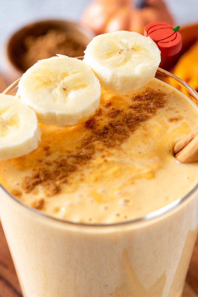 A thick and creamy blended pumpkin smoothie in a glass that's garnished with three slices of banana on a pumpkin toothpick stick and pumpkin pie spice sprinkled on top.
