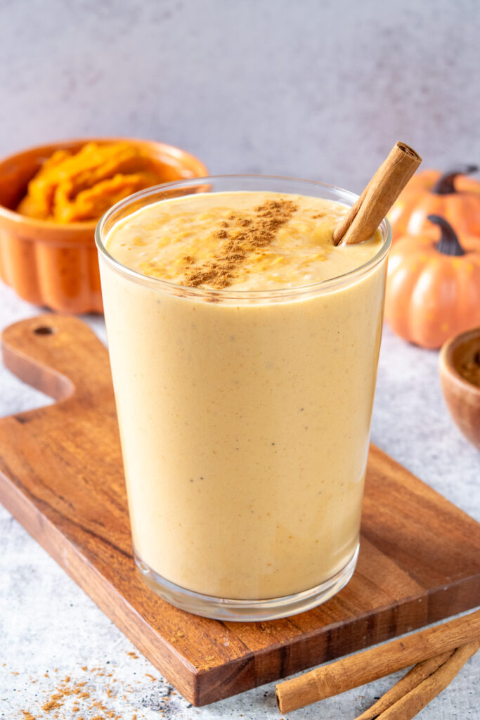 A homemade pumpkin smoothie in a glass with cinnamon sprinkled on top. In the background are some mini pumpkins and an orange bowl filled with pumpkin puree.