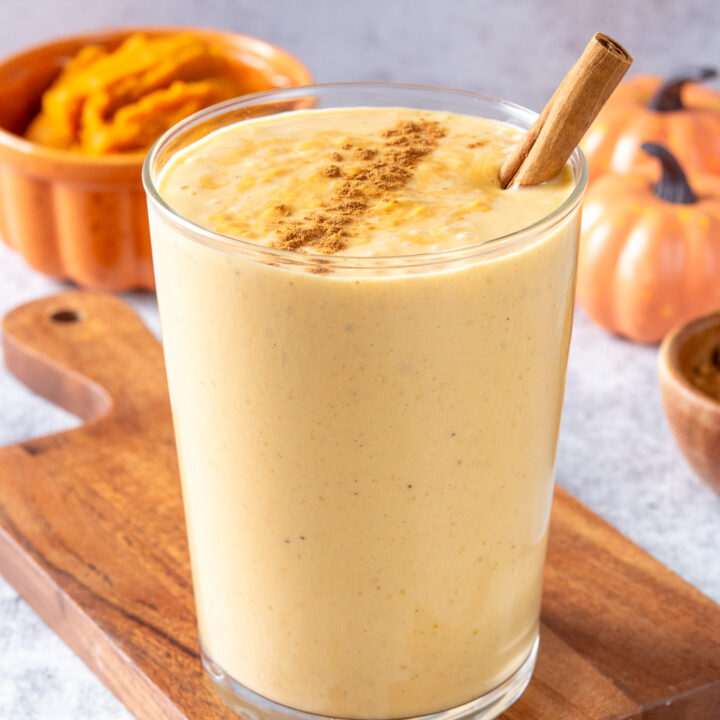 A homemade pumpkin smoothie in a glass with cinnamon sprinkled on top. In the background are some mini pumpkins and an orange bowl filled with pumpkin puree.