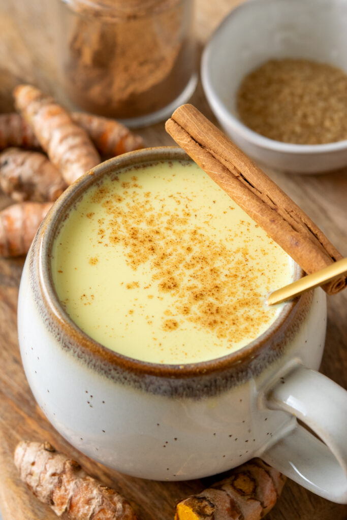 A close-up view of a homemade turmeric latte with cinnamon sprinkled on top, showing it's golden color. There's a cinnamon stick resting on the edge of the latte and some turmeric roots scattered around.