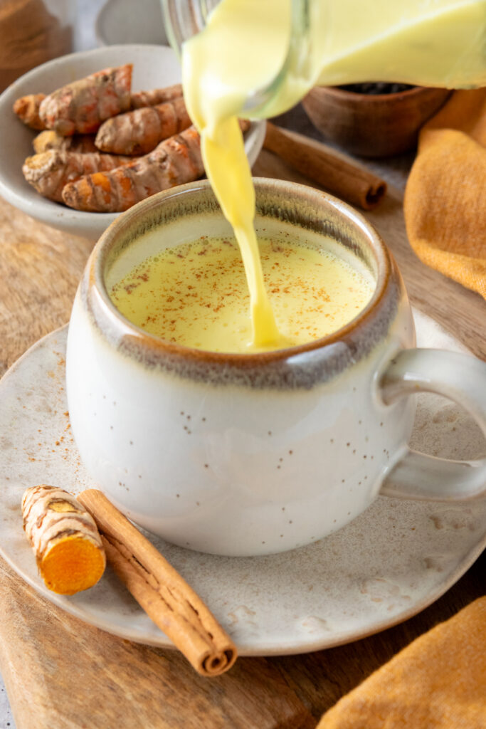 Pouring yellow-colored golden milk into a mug. There's sliced turmeric root next to the mug and a cinnamon stick.