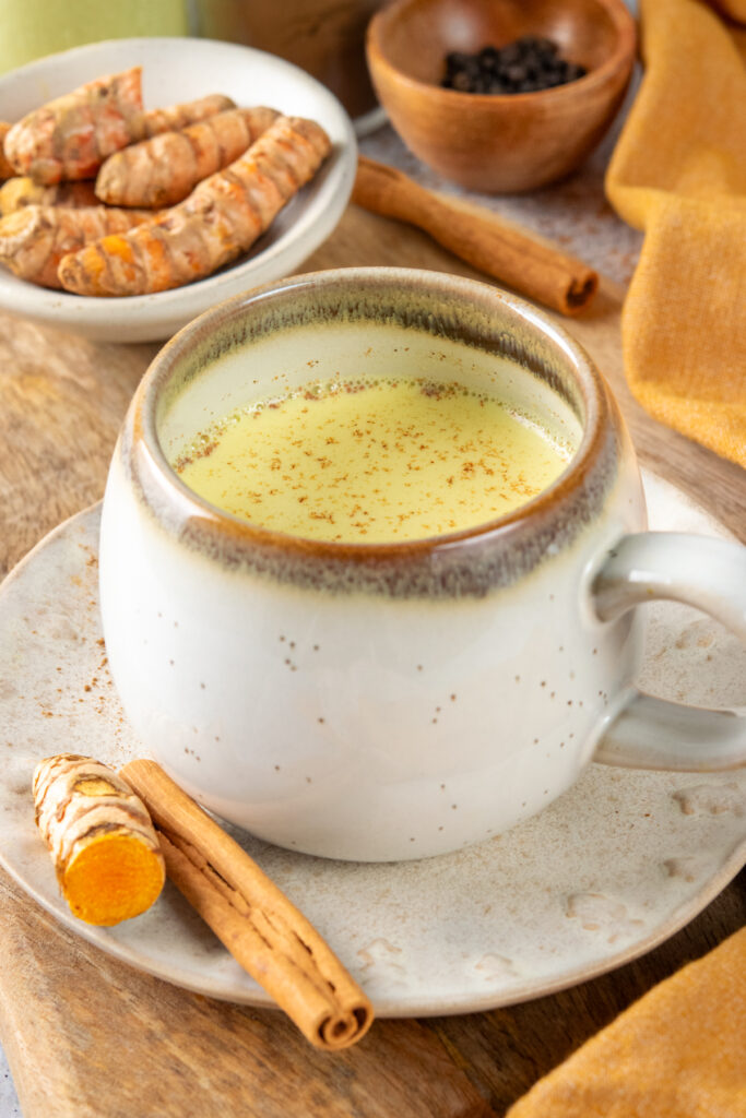 A turmeric latte (golden milk) in a mug. There's sliced turmeric root next to the mug and in a small bowl of turmeric root and cinnamon sticks in the background.