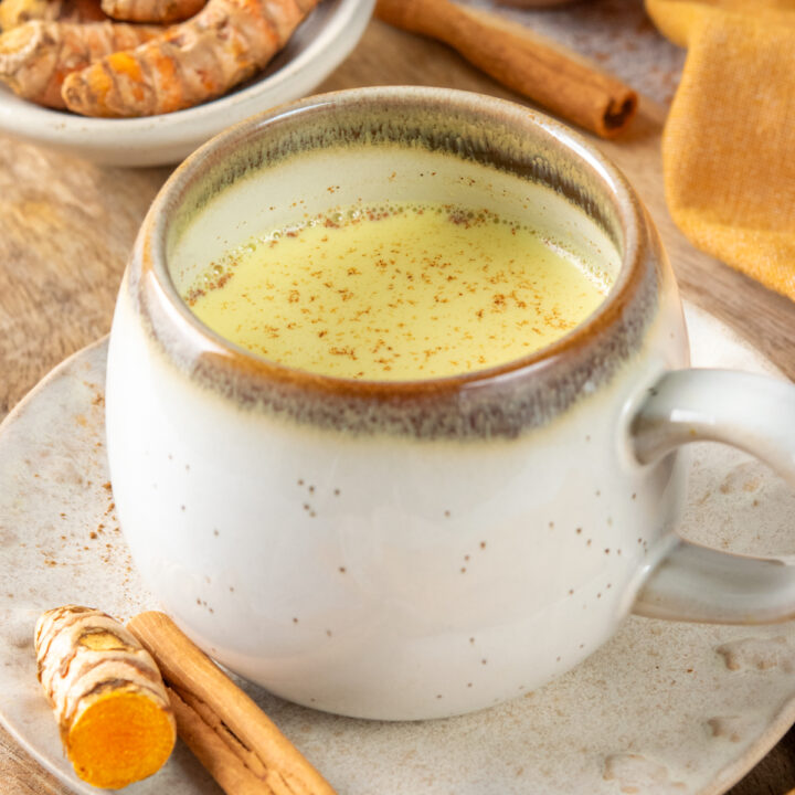 A turmeric latte (golden milk) in a mug. There's sliced turmeric root next to the mug and in a small bowl of turmeric root and cinnamon sticks in the background.