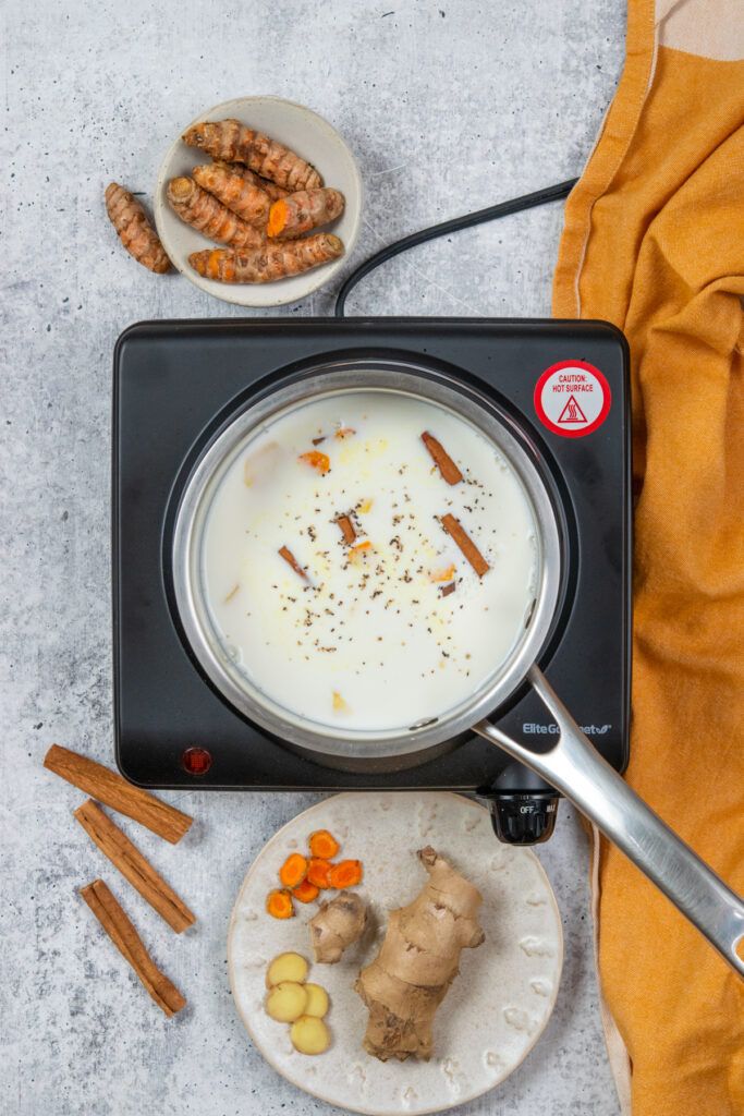 A saucepan with milk, fresh turmeric, fresh ginger, cinnamon sticks and black pepper.
