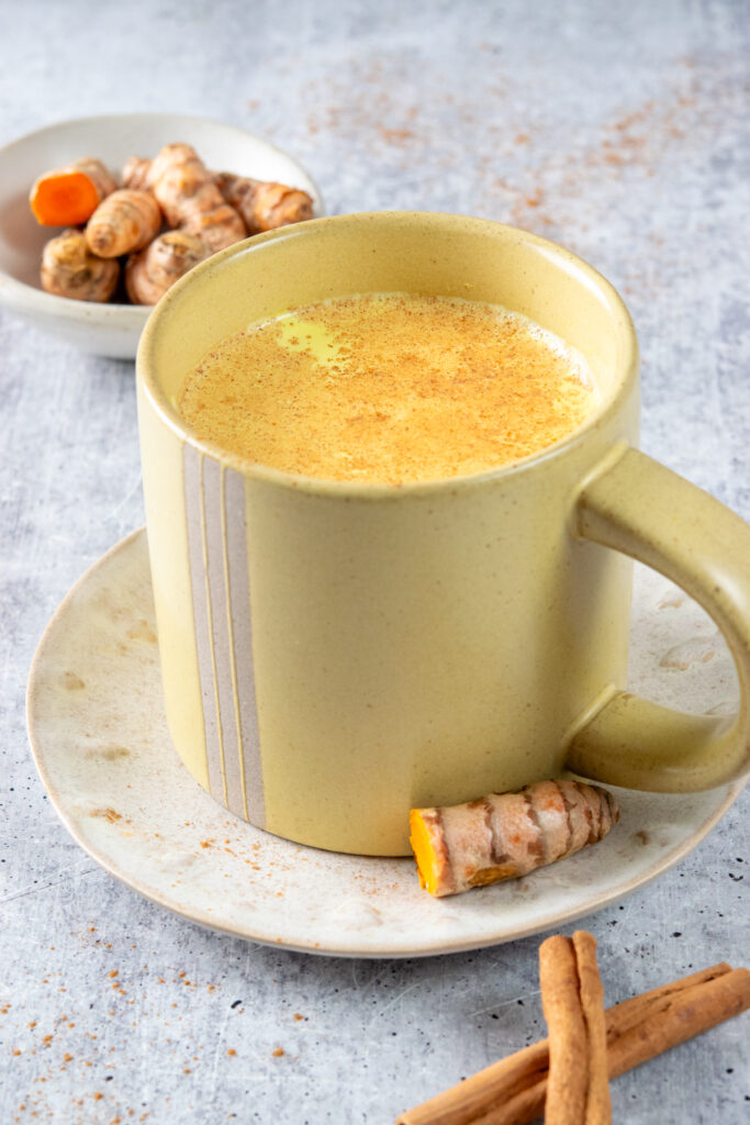 A golden milk latte that has cinnamon sprinkled on top, in a yellow mug. Behind the latte is a small bowl filled with turmeric root. One of the pieces is cut in half showing how orange it is.