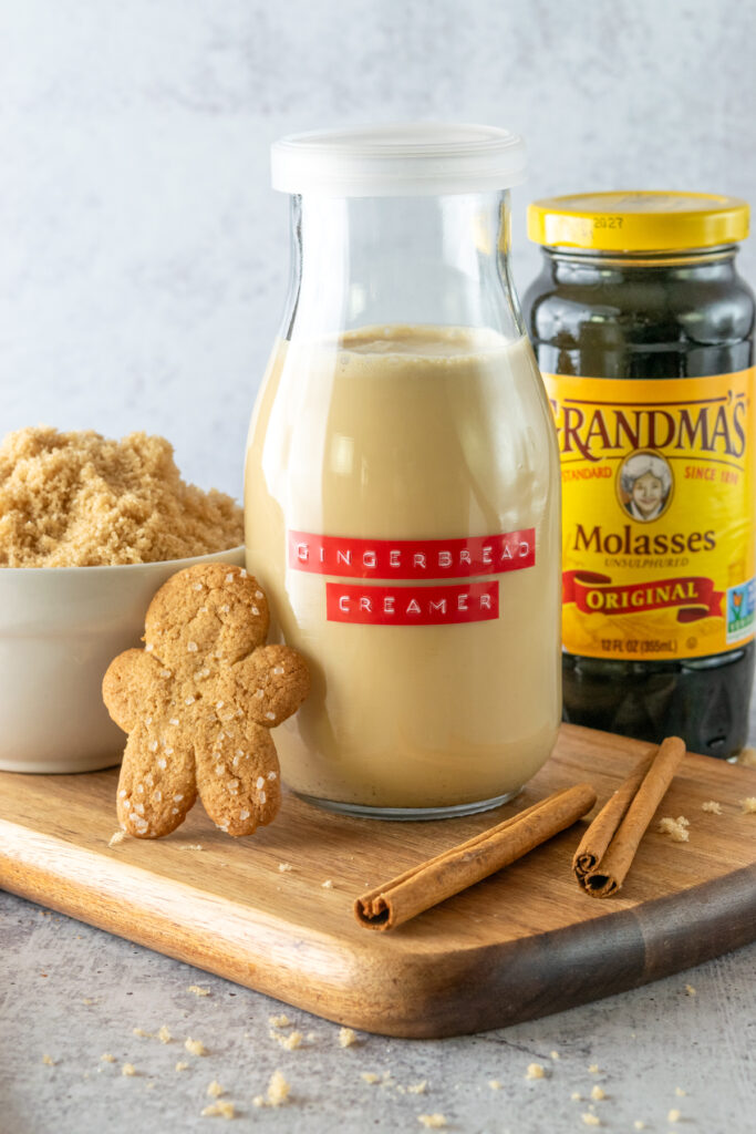 Small bottle of homemade coffee creamer labeled, "gingerbread creamer" in front of a jar of molasses, bowl of brown sugar and gingerbread cookie.