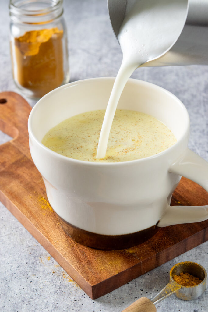 Pouring thick steamed milk into a latte mug that has turmeric powder mix in it. In front of the mug is a measuring spoon that was used to scoop the turmeric latte mix out of the jar. The latte is golden and frothy.
