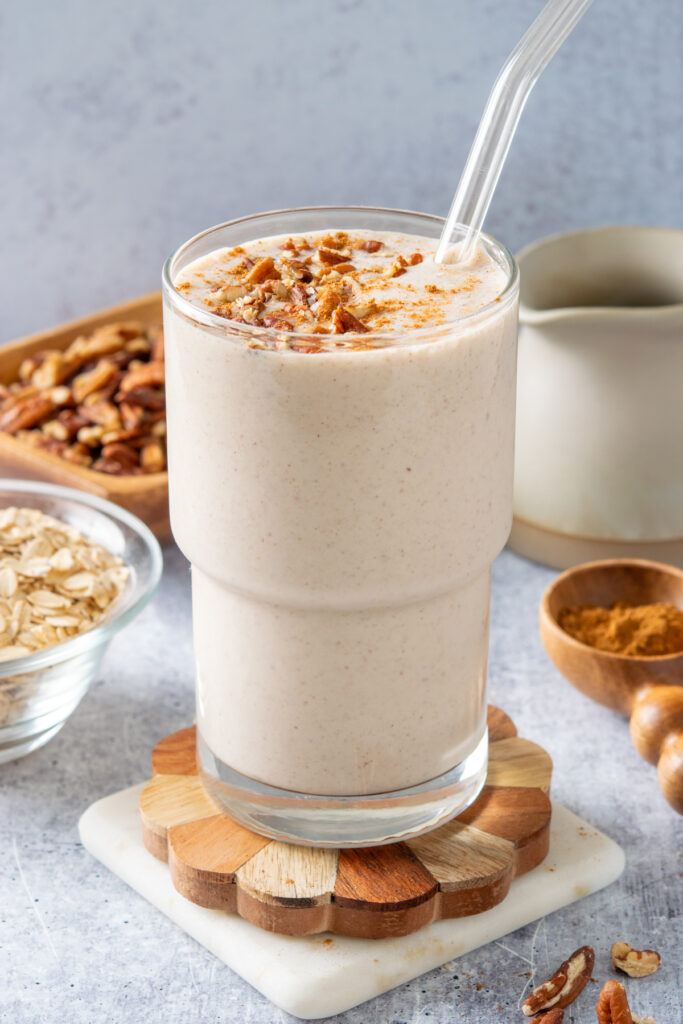 A maple pecan oatmeal breakfast smoothie surrounded by bowl of oats, bowl of pecans, pitcher of maple syrup, and a measuring spoon filled with ground cinnamon.
