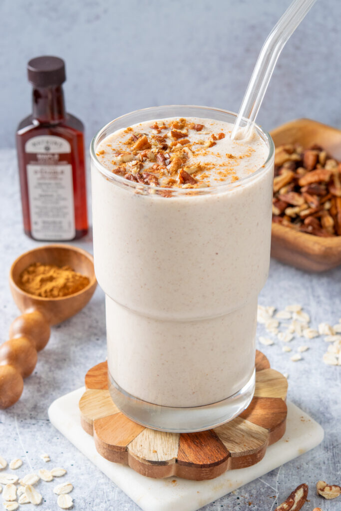 A pecan oatmeal smoothie topped with pecans and cinnamon in a glass which is next to a bottle of maple extract, a wood spoon with ground cinnamon, and a small wooden bowl filled with pecans. There are oats sprinkled around the smoothie on the table.