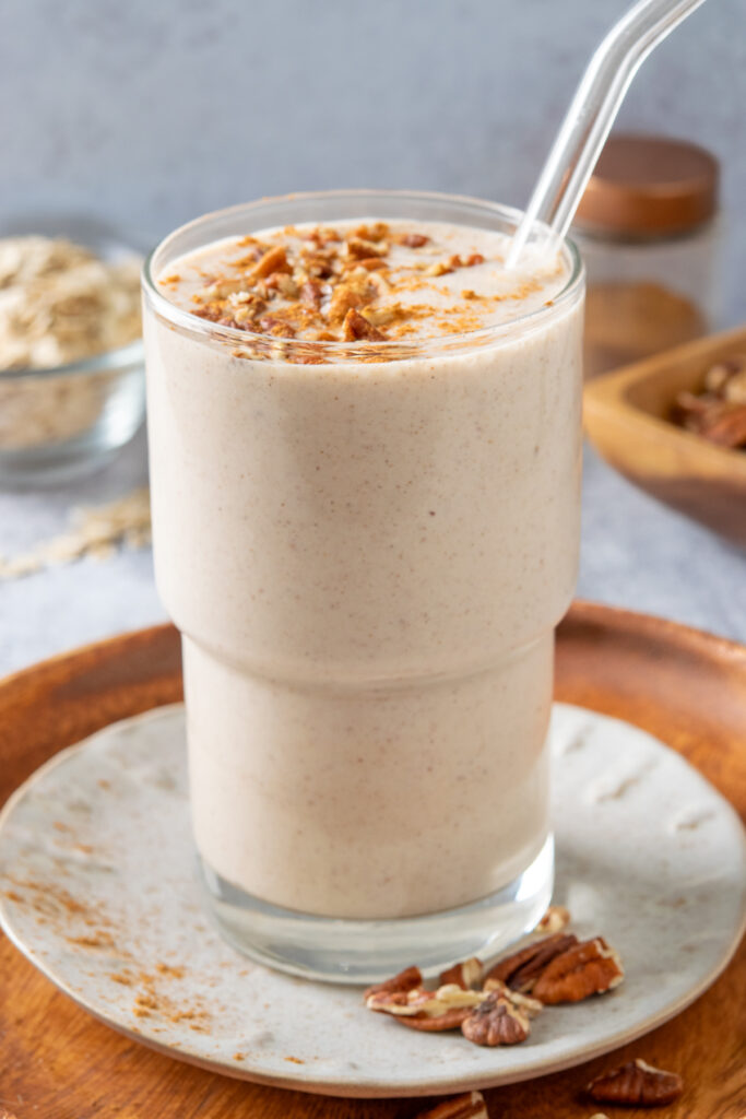 A glass filled with a maple-flavored oatmeal smoothie that has chopped pecans and cinnamon on top. The smoothie is sitting on a plate which is on a wooden tray.