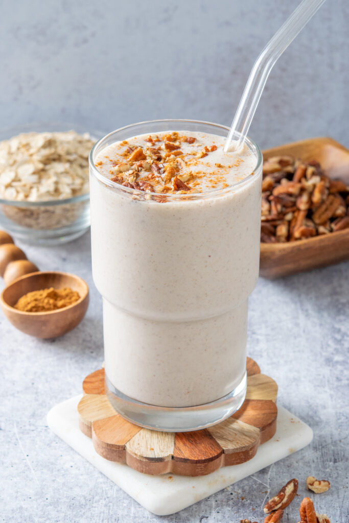 A homemade maple pecan oatmeal smoothie that's garnished with bits of pecan and cinnamon. Behind the smoothie is a bowl of oats and a bowl of pecans.