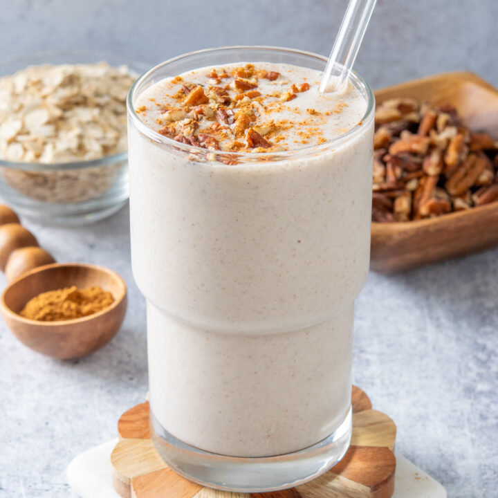 A homemade maple pecan oatmeal smoothie that's garnished with bits of pecan and cinnamon. Behind the smoothie is a bowl of oats and a bowl of pecans.