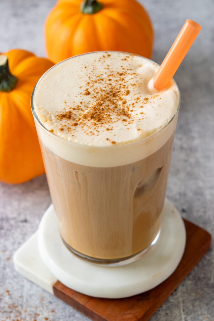 Homemade pumpkin cold foam topping an iced cold brew drink. There is pumpkin spice sprinkled on top of the creamy cold foam, there are two pumpkins behind the drink which also has an orange straw in it.