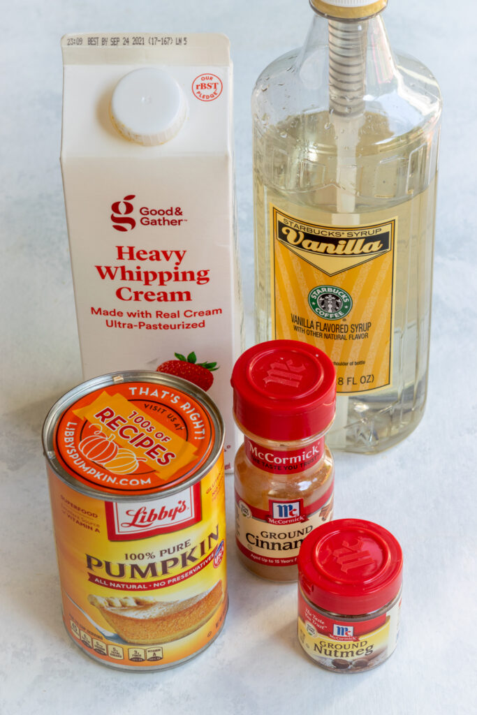 Ingredients in packages needed to make Starbucks copycat pumpkin cold foam, including a carton of heavy cream, can of pumpkin puree, bottle of Starbucks vanilla syrup, and spice jars with ground cinnamon and ground nutmeg.