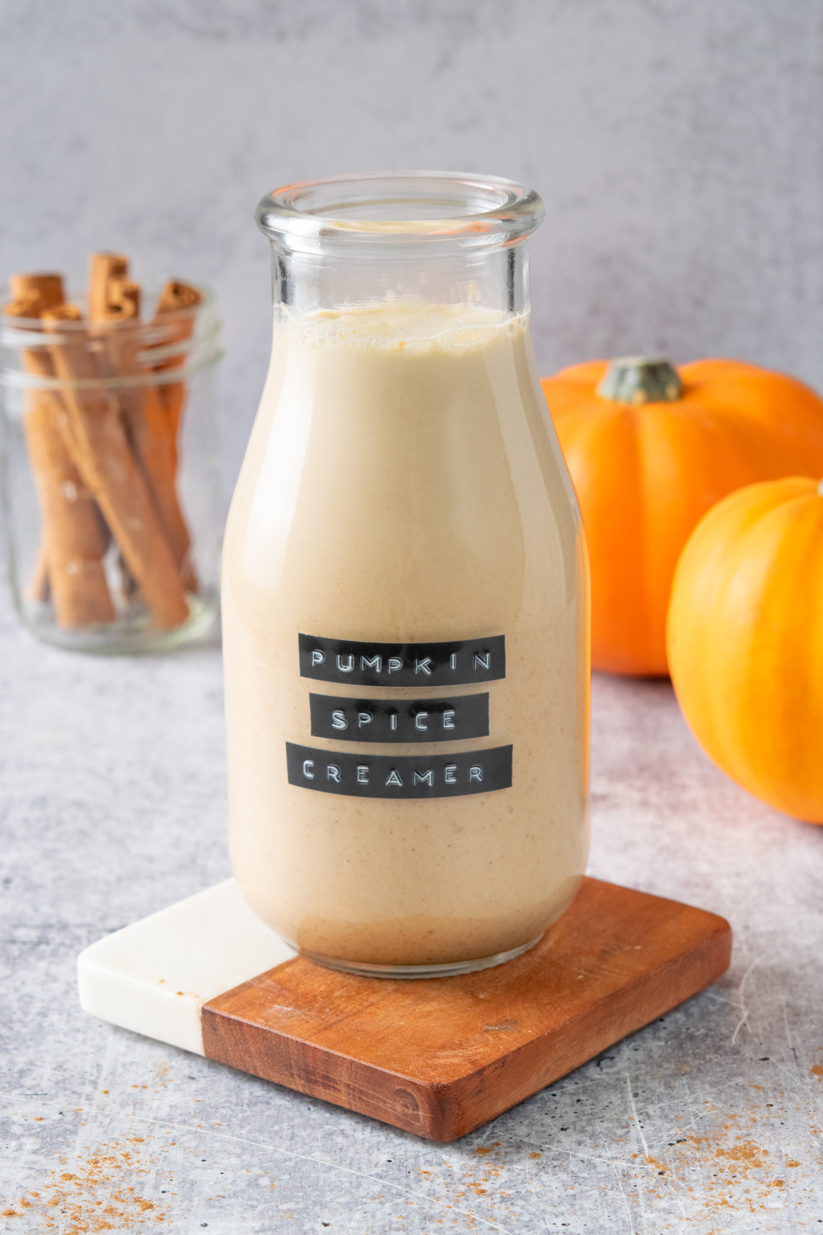 Homemade pumpkin spice coffee creamer in a bottle with a label. There are two pumpkins and a jar of cinnamon sticks in the background.