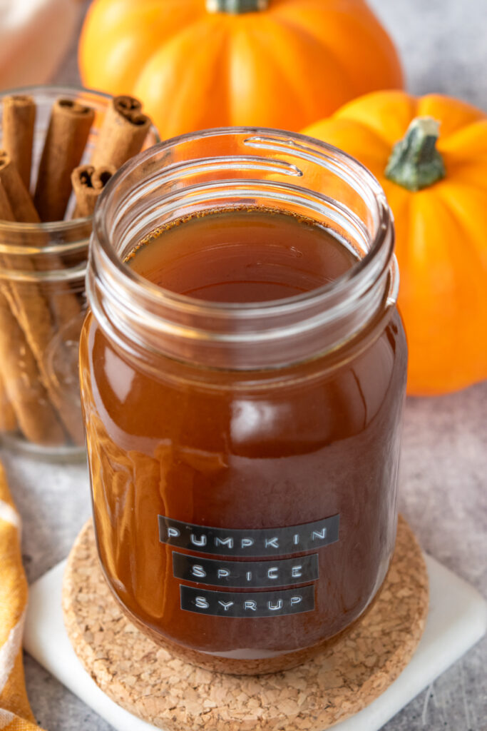 Homemade pumpkin spice syrup in a jar which is next to some cinnamon sticks and pumpkins. The jar is labeled, "pumpkin spice syrup."