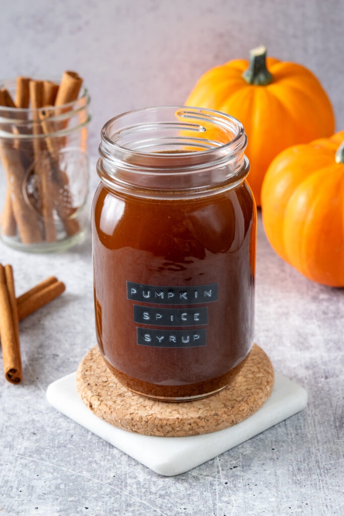 A mason jar that has a "pumpkin spice syrup" label on it and is filled with the homemade coffee sryup. Next to the syrup are a few cinnamon sticks and mini pumpkins.