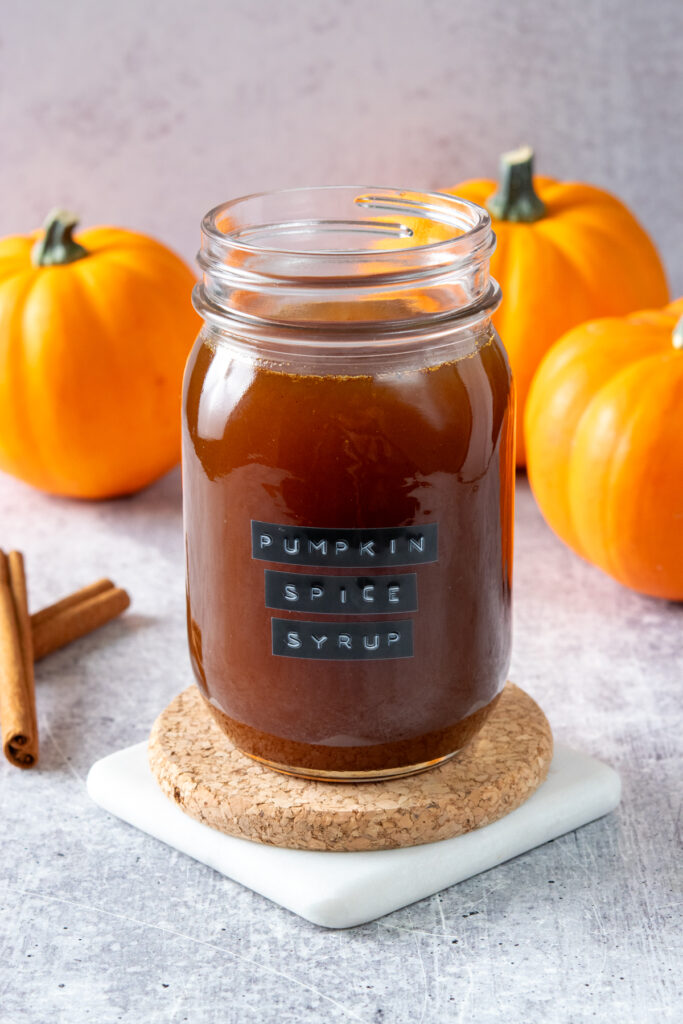 Homemade pumpkin spice syrup in a jar with a "pumpkin spice syrup" label. There are a few pumpkins and cinnamon sticks behind the latte syrup.