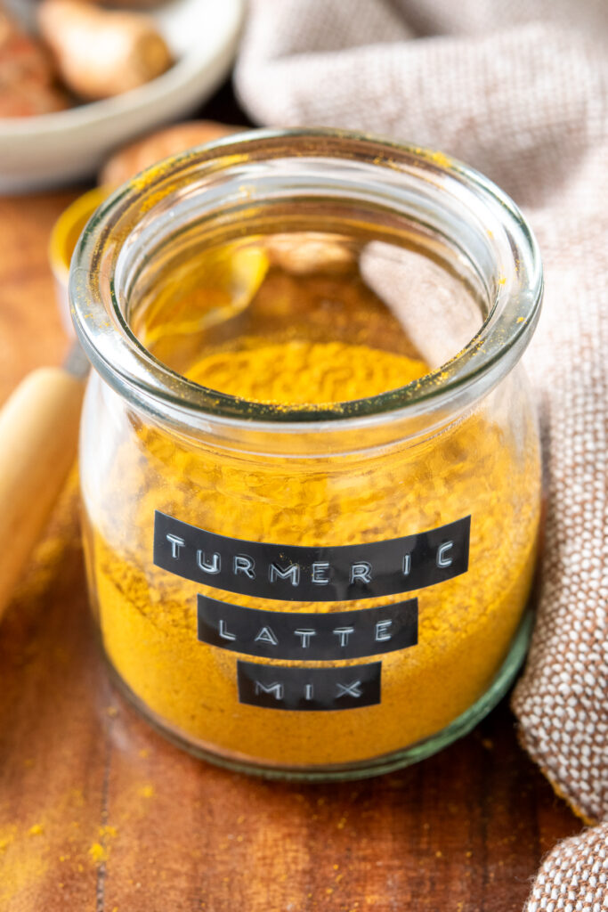 A small jar of homemade turmeric latte mix. The jar has a labeled, "turmeric latte mix." There is a scoop with some turmeric powder on it and a small bowl of turmeric root in the background.