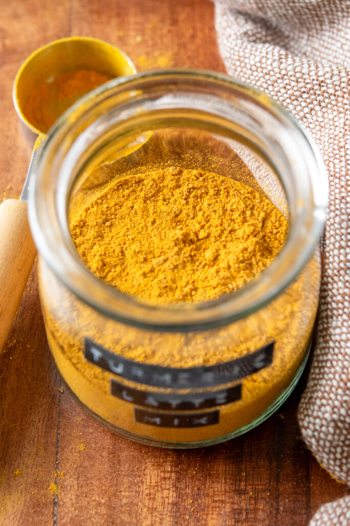 A close-up view looking into the top of a jar of turmeric latte mix. The mix is a deep golden color. Next to the jar is a measuring spoon that has some of the  powder on it.